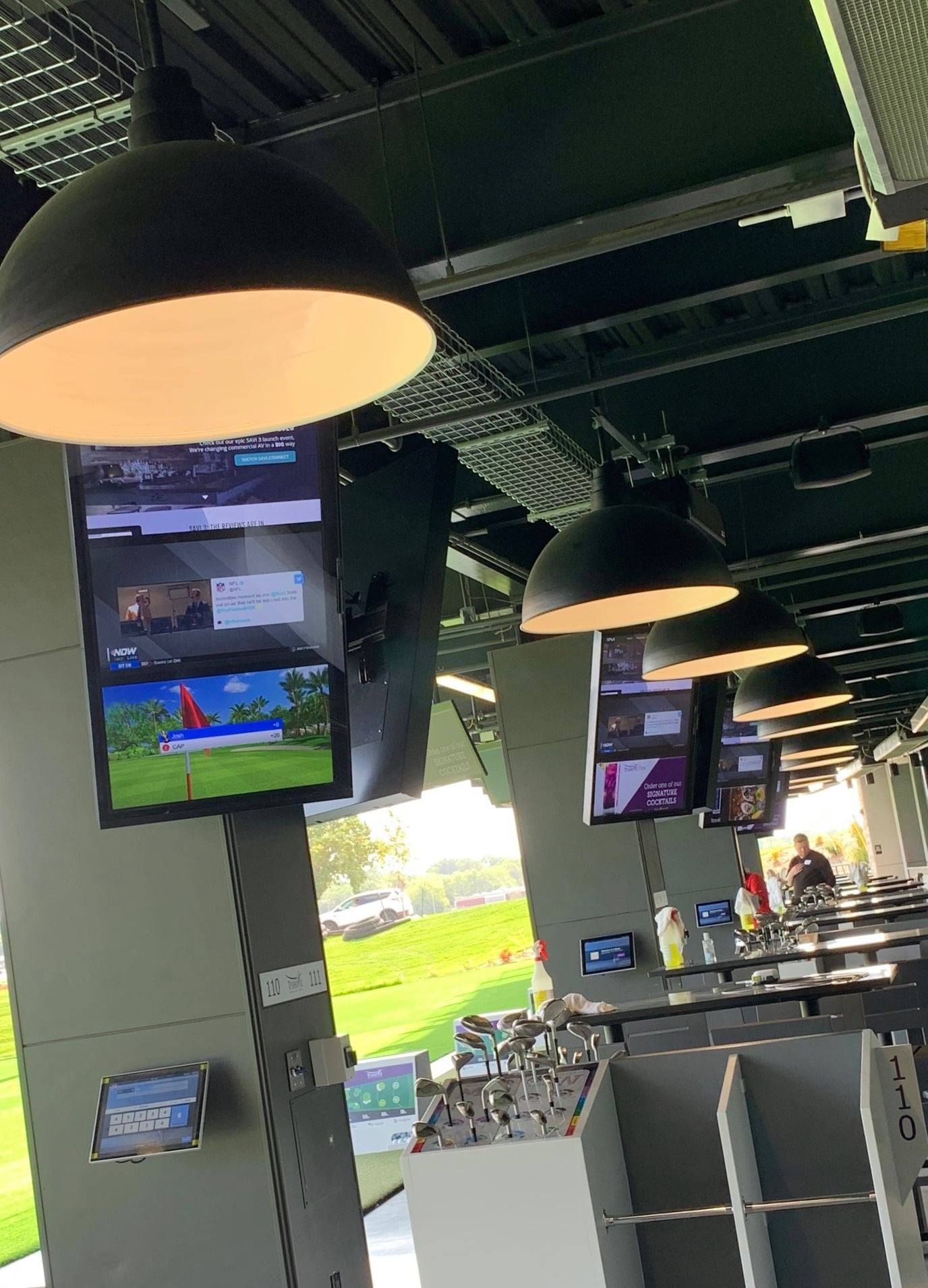 Golf driving range with screens, hanging lights, and a grassy outdoor view.