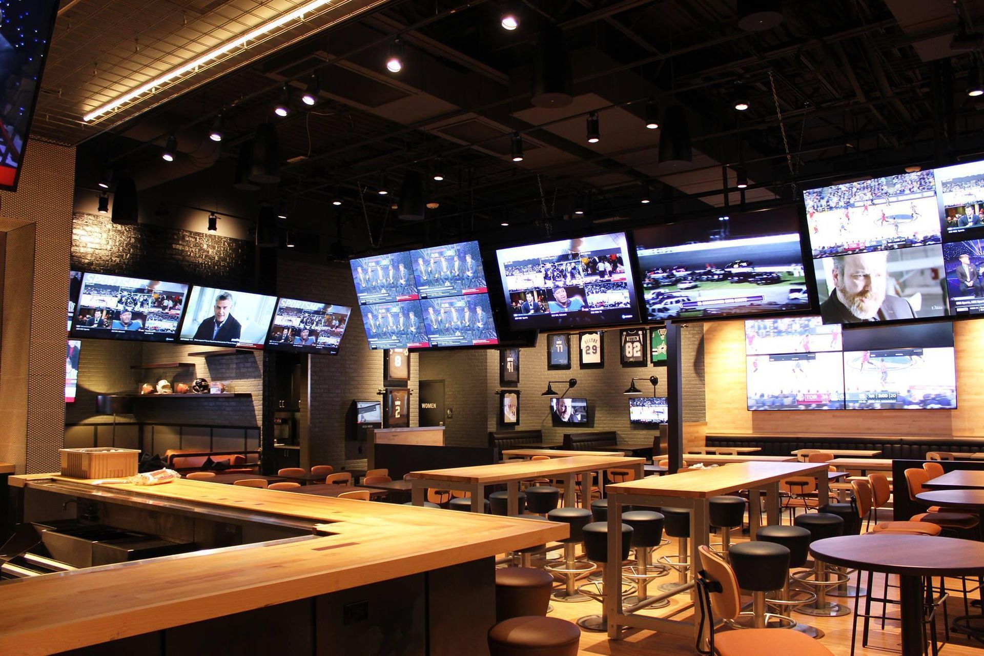 Interior of a sports bar with many TVs displaying various games. Wooden tables, bar stools, and a bar area are visible.