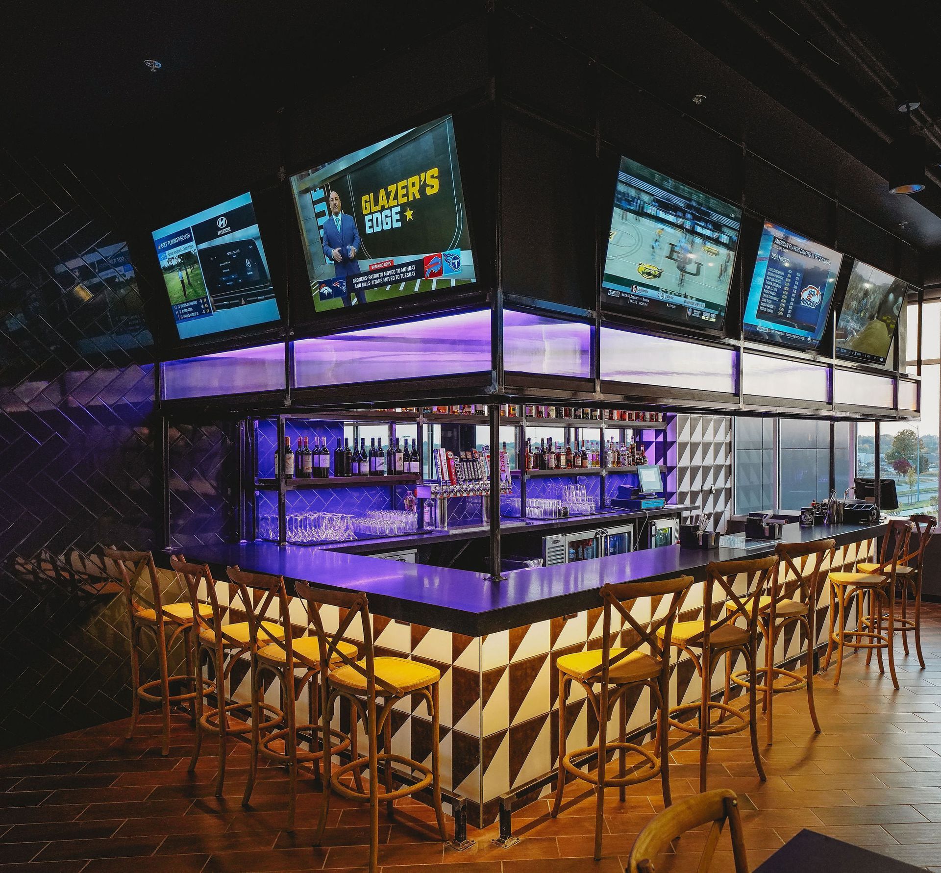 Bar with multiple TVs showing sports. Yellow chairs, purple lighting, and bottles on shelves.