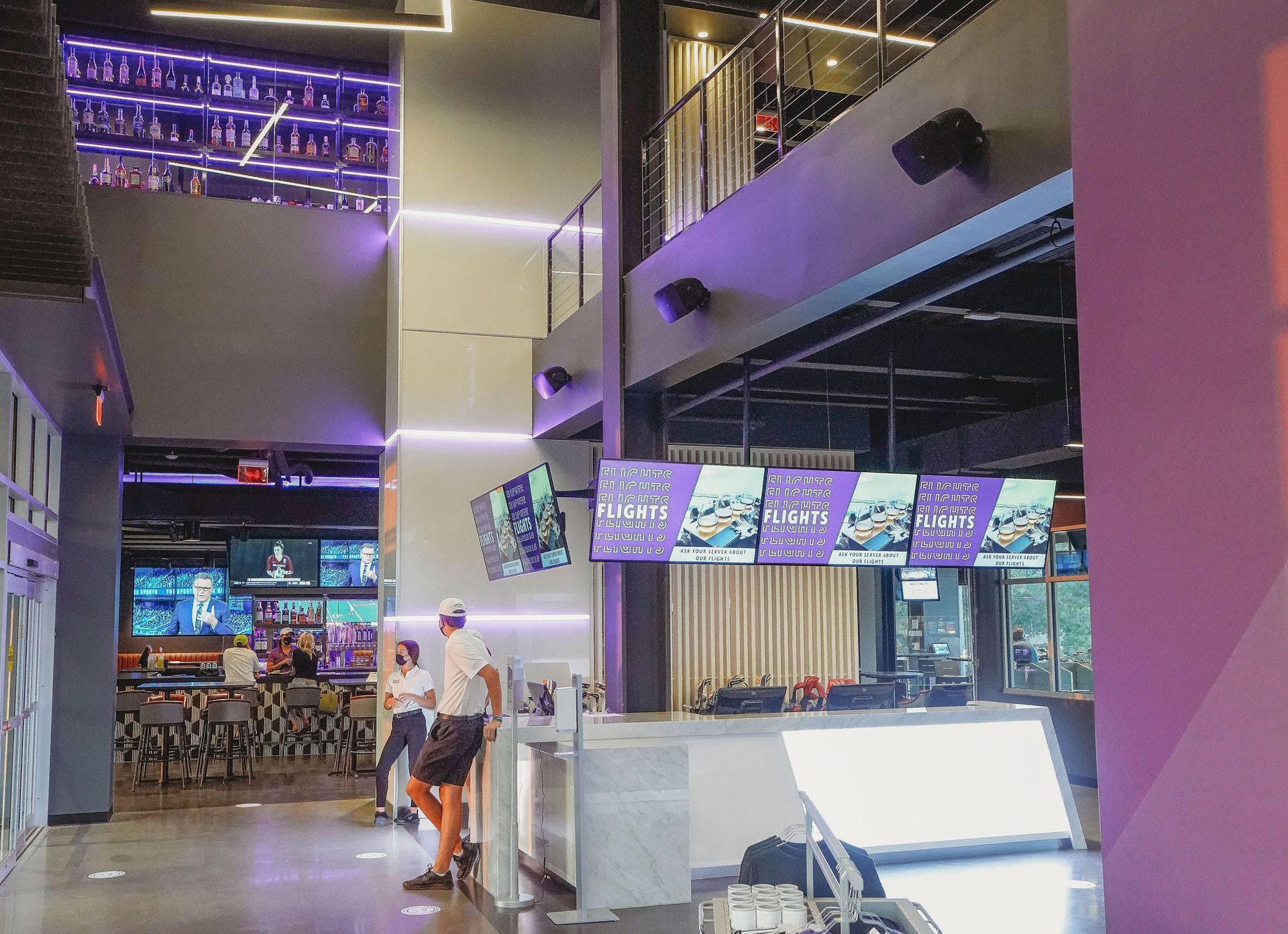 Interior of a modern sports bar. Two-story with screens, purple accents, people standing.