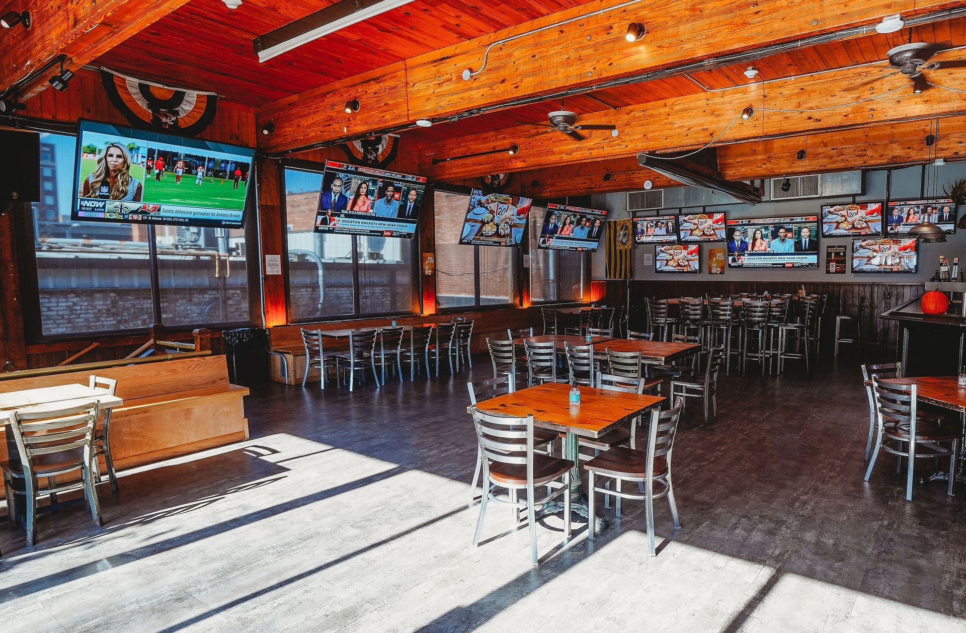 Interior of a sports bar with many TVs, tables, and chairs. Sunlight streams in. Wooden ceiling.