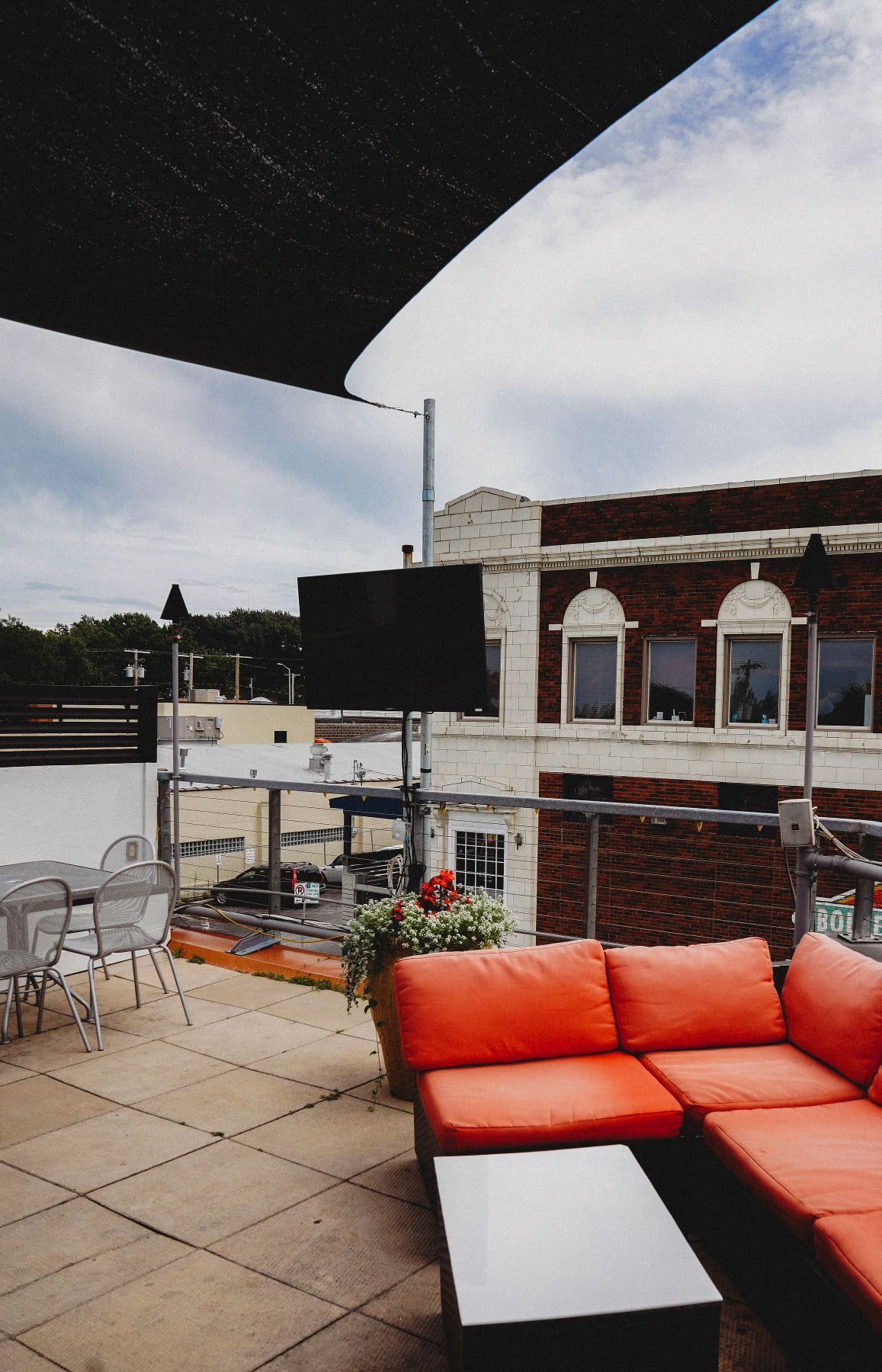 Rooftop patio with orange sectional sofa, black shade, TV, and brick building background under cloudy sky.