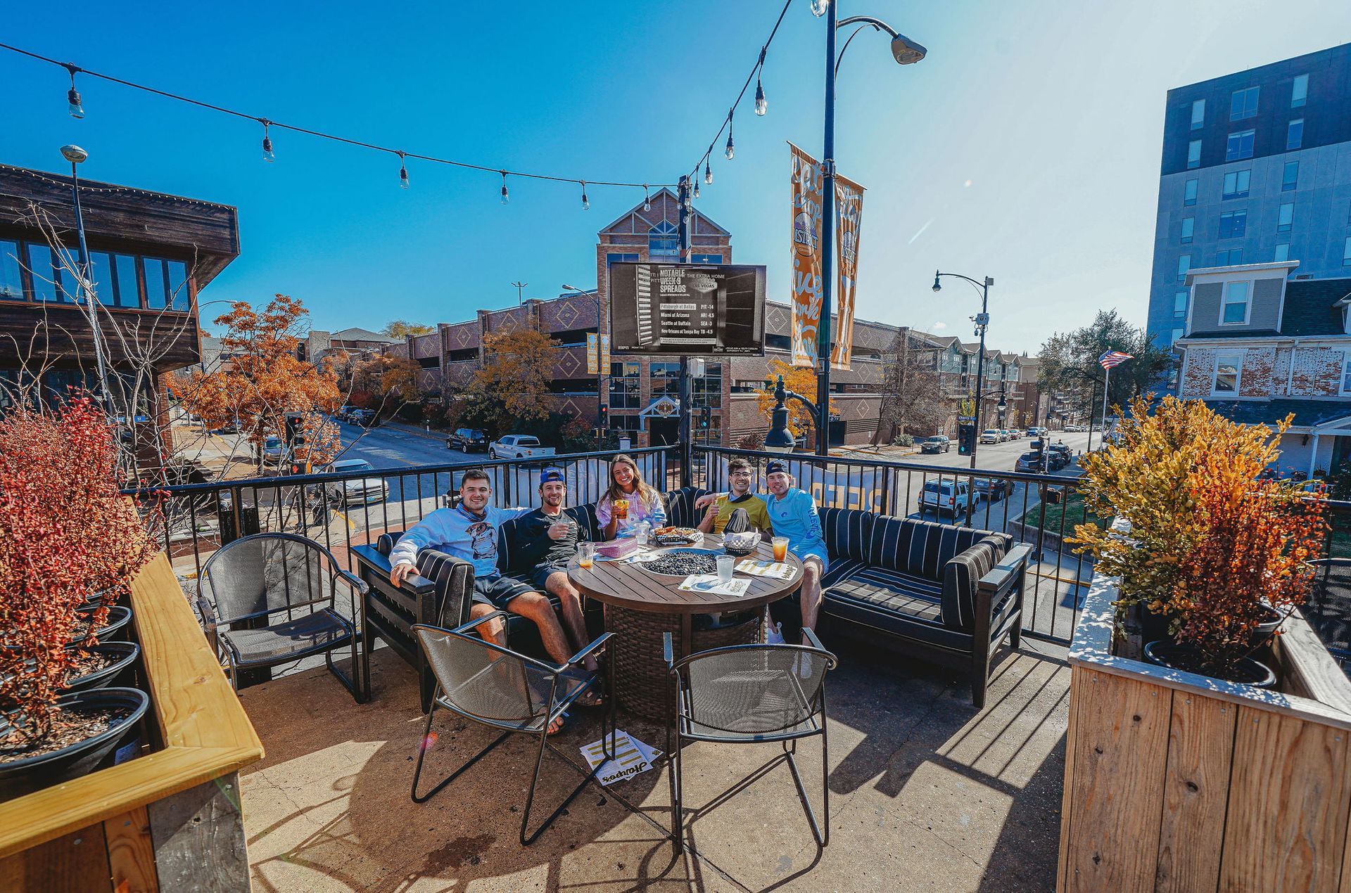 Friends on a sunny patio, seated around a wooden table with food and drinks. City buildings in background.
