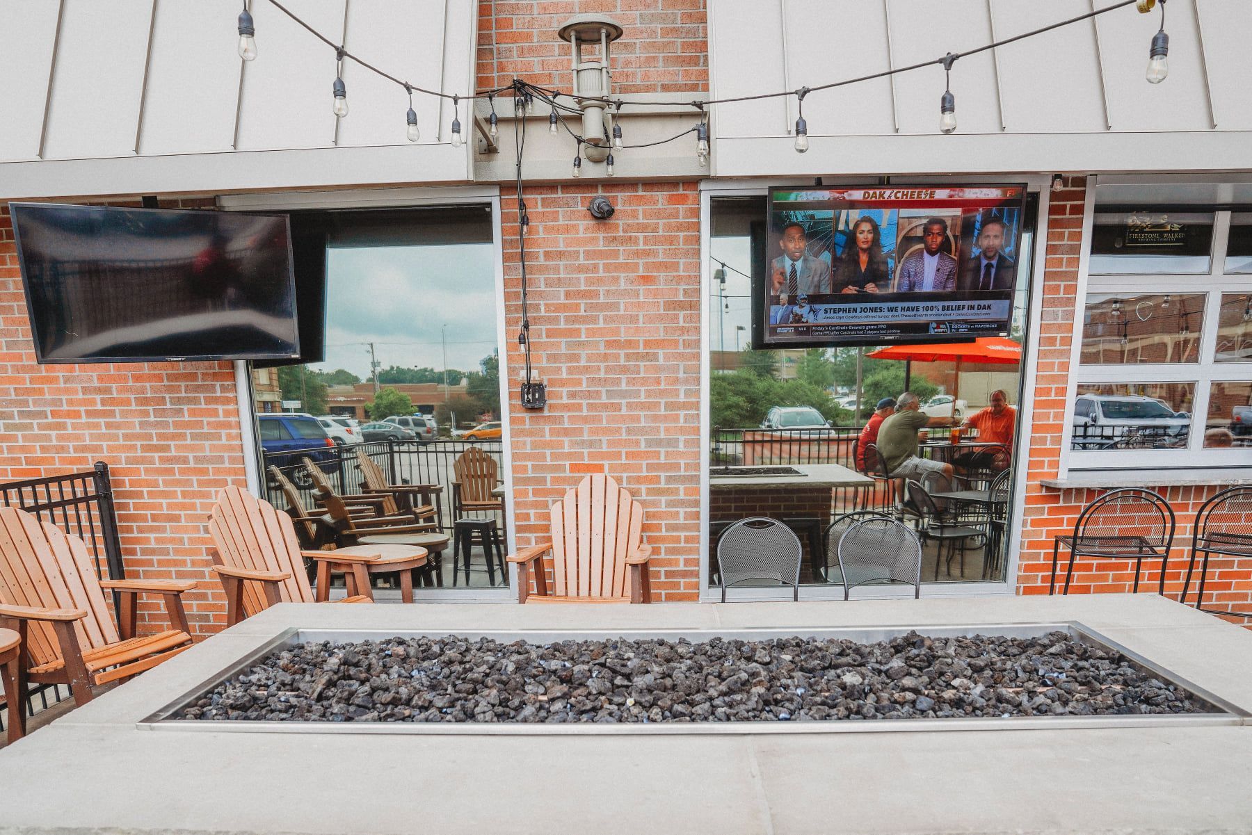 Outdoor patio with fire pit, TVs, and seating near a brick building. People visible inside, watching TV.