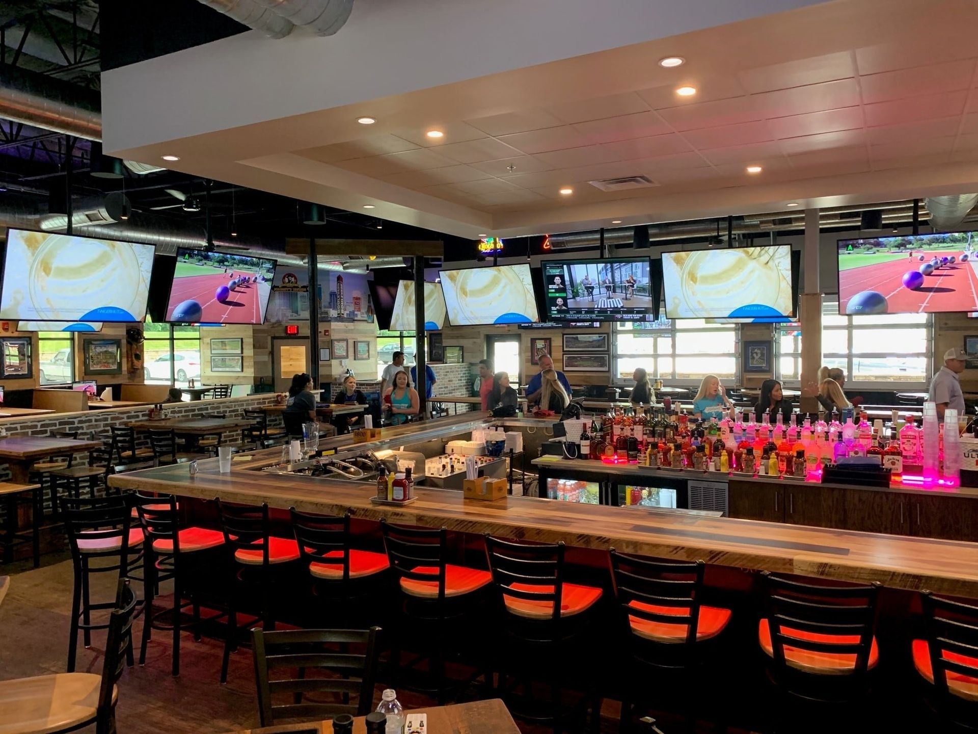Bar interior with multiple TVs showing sports, barstools, and people seated at tables, lit with red and white lights.