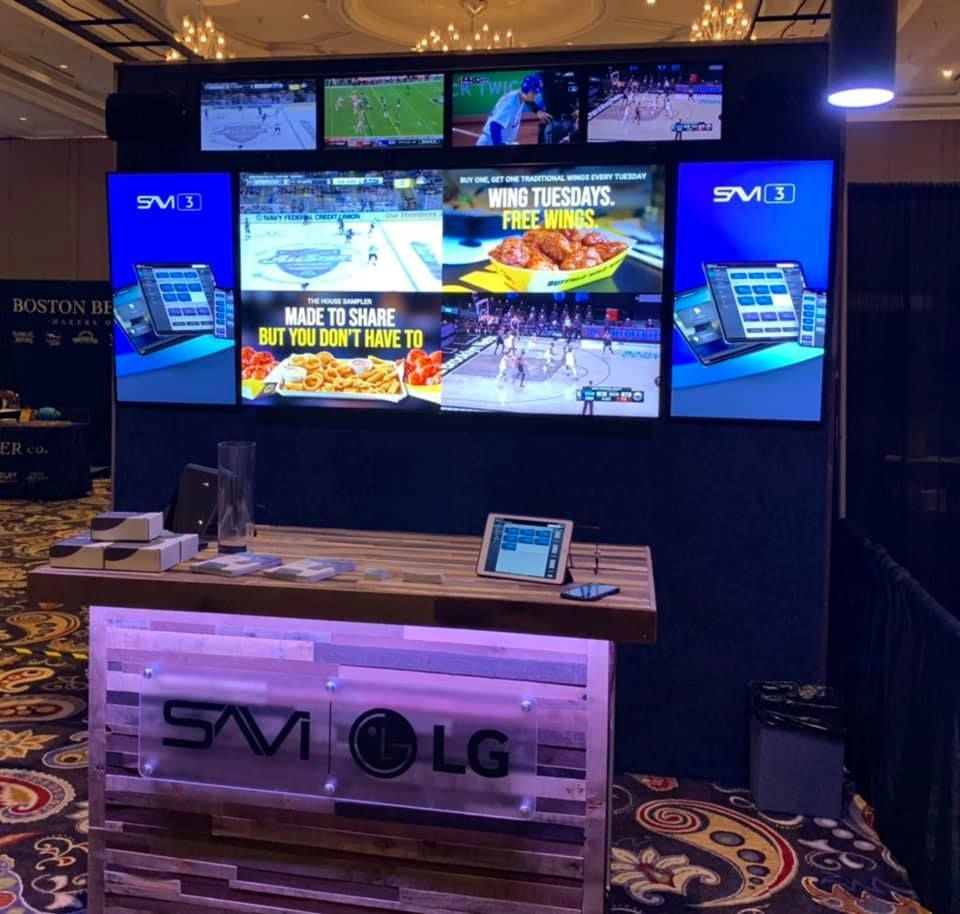 Trade show booth with TVs displaying sports and food ads; bar with tablets and logo.