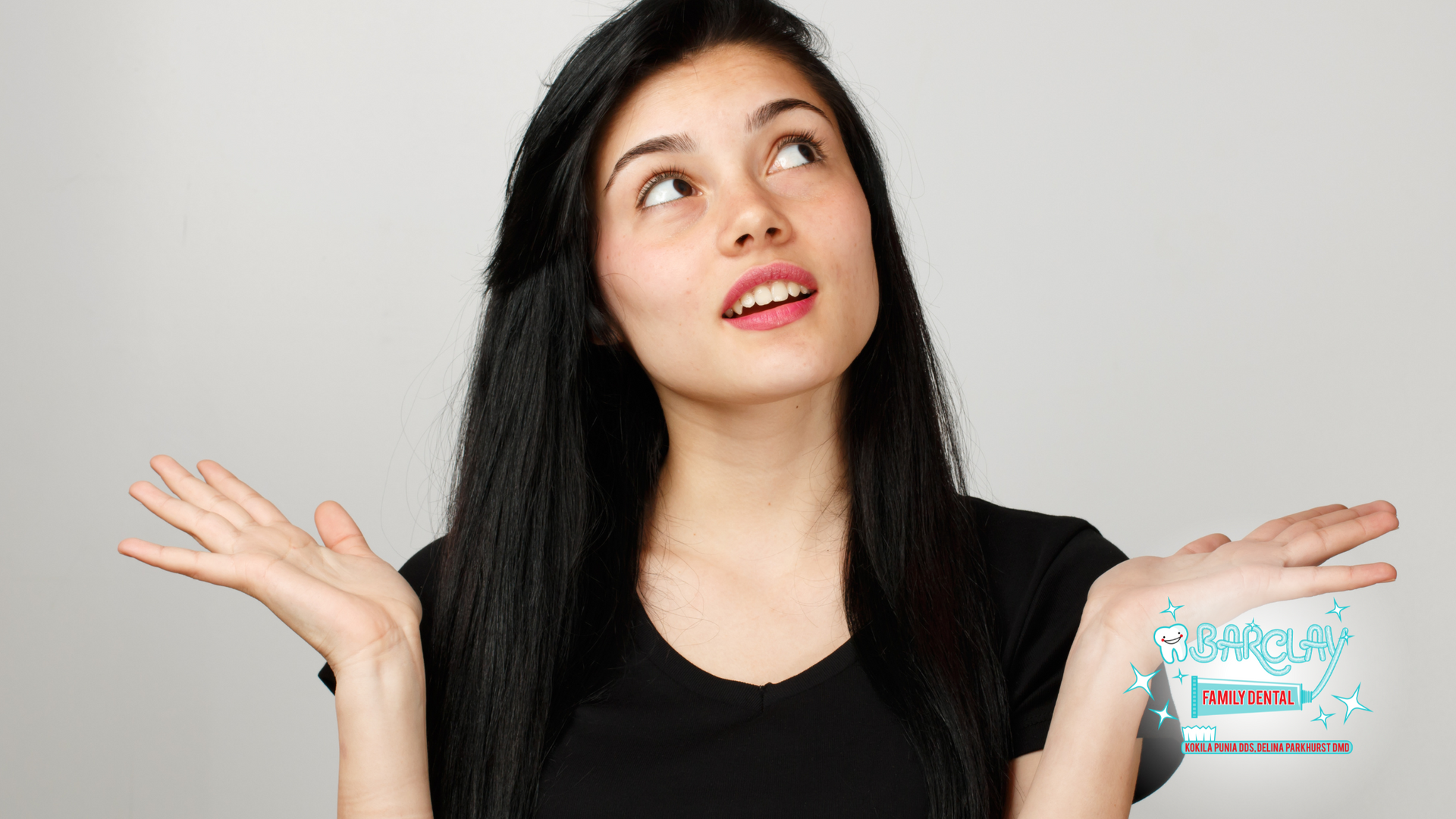 Woman looking up, shrugging with open palms, wearing a black shirt, against a gray backdrop.