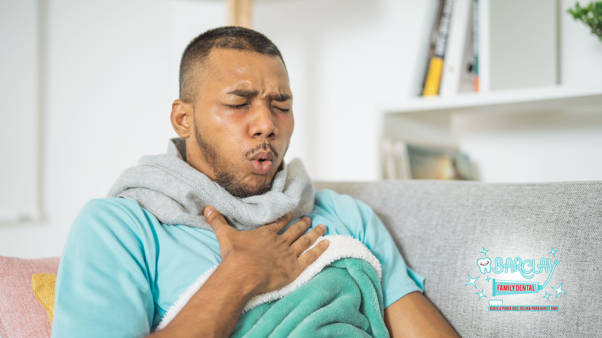 A man is sitting on a couch with a blanket around his neck and coughing.