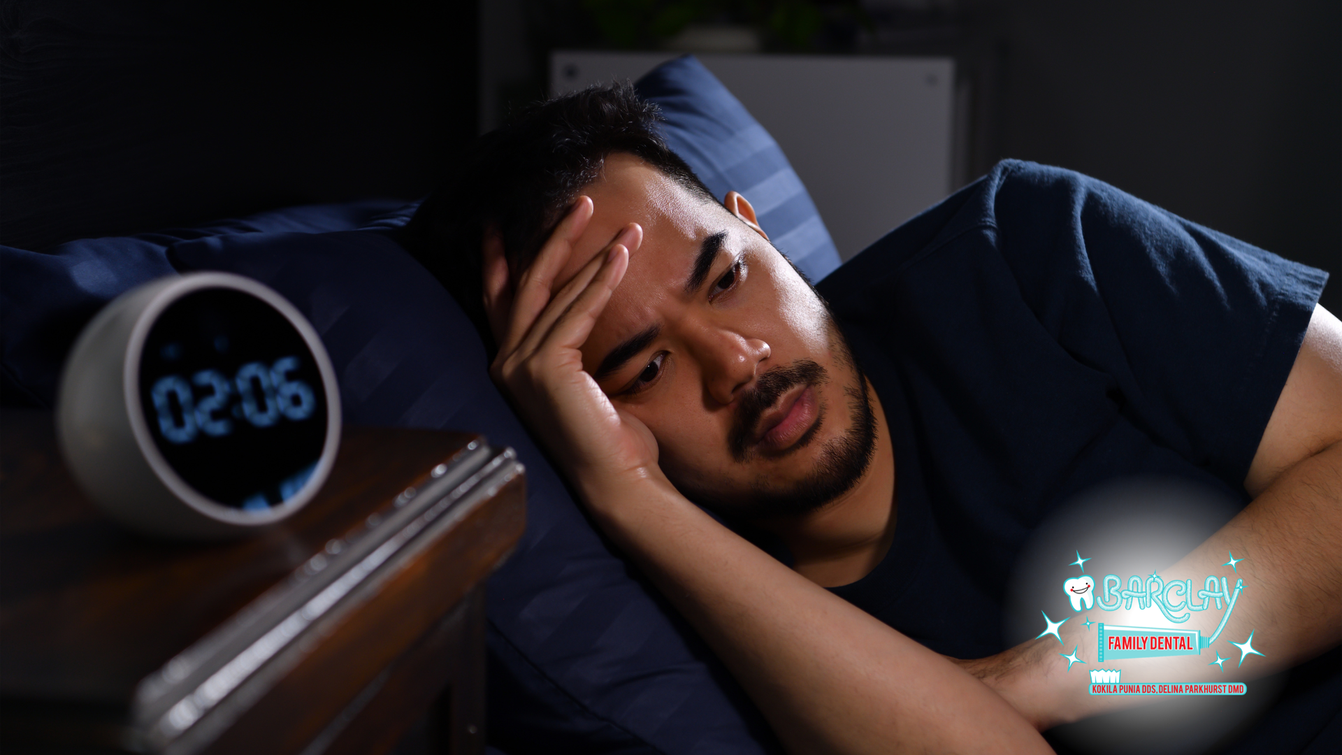 Man lying in bed with hand on forehead beside a glowing digital alarm clock, looking tired in dim light