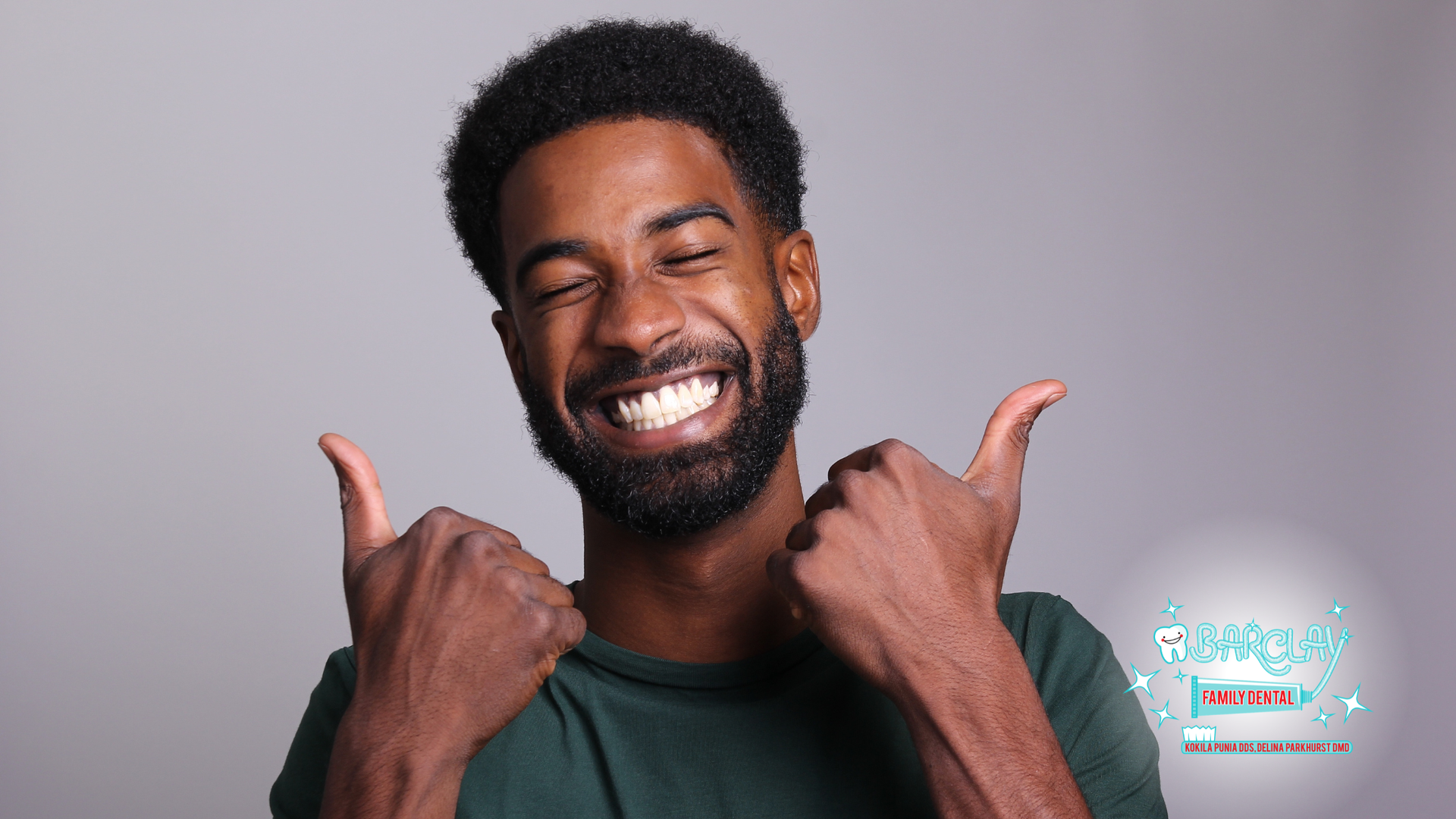 A smiling person wearing a dark green shirt gives a thumbs-up with both hands against a light gray background.