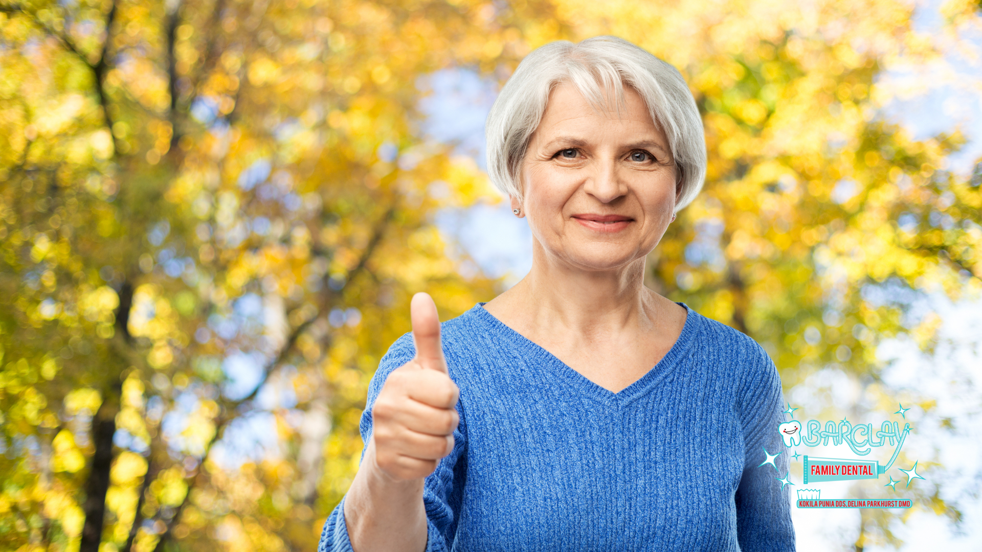 An elderly woman in a blue sweater is giving a thumbs up.