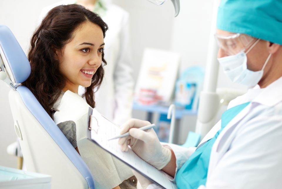 Woman in a dental chair smiles as dentist, wearing mask and gloves, reviews a chart.