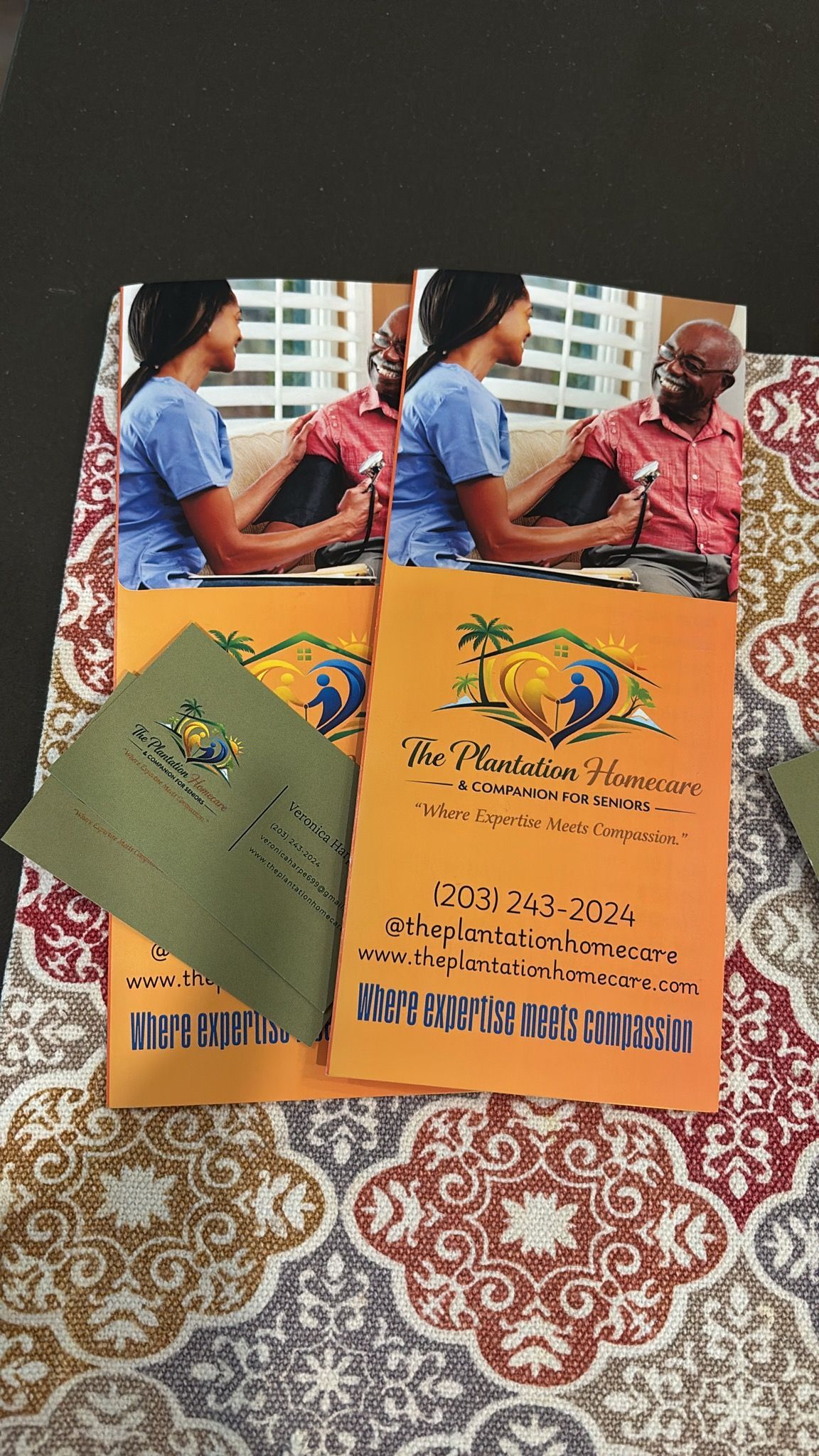 Two orange event flyers and a green booklet on a patterned cloth, with a person holding one flyer.