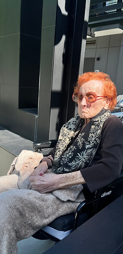 Person with orange hair and sunglasses sitting in a wheelchair outdoors, holding a blanket in a black coat.