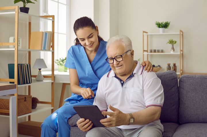 Caregiver standing beside a man on a sofa, both looking at a tablet in a bright living room