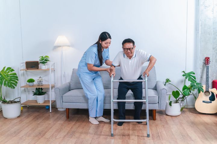 Woman assisting man with walker in a bright living room with couch, lamp, and plants