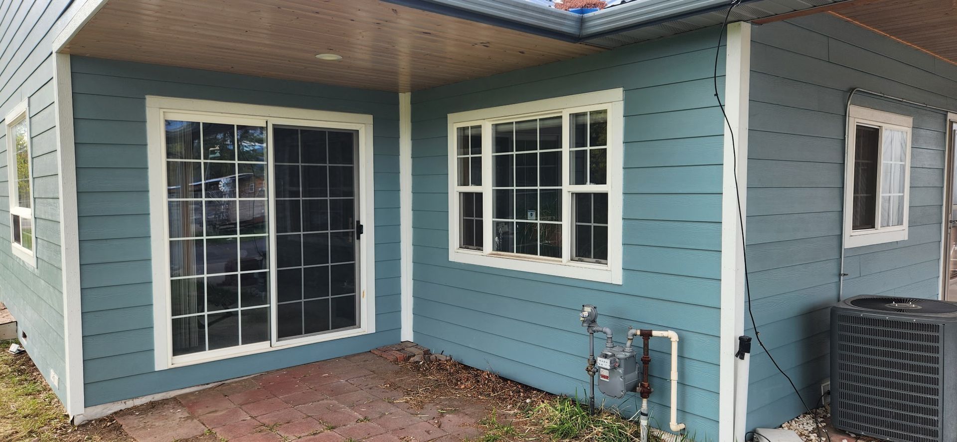 Blue-sided house with white-trimmed windows and sliding glass door. A/C unit on the side. Brown patio.