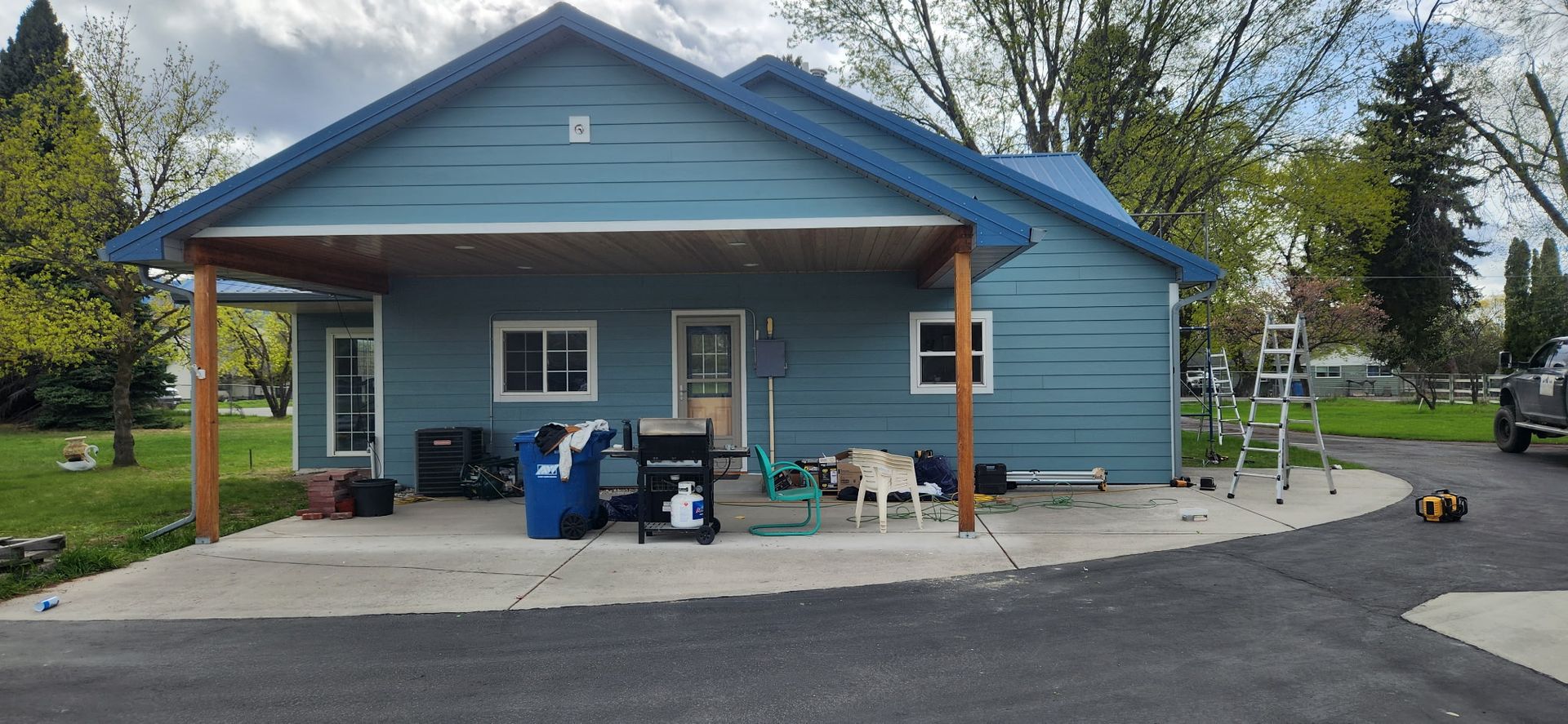 Blue house with a carport, yard, and driveway. Items on concrete. Ladder by the side.