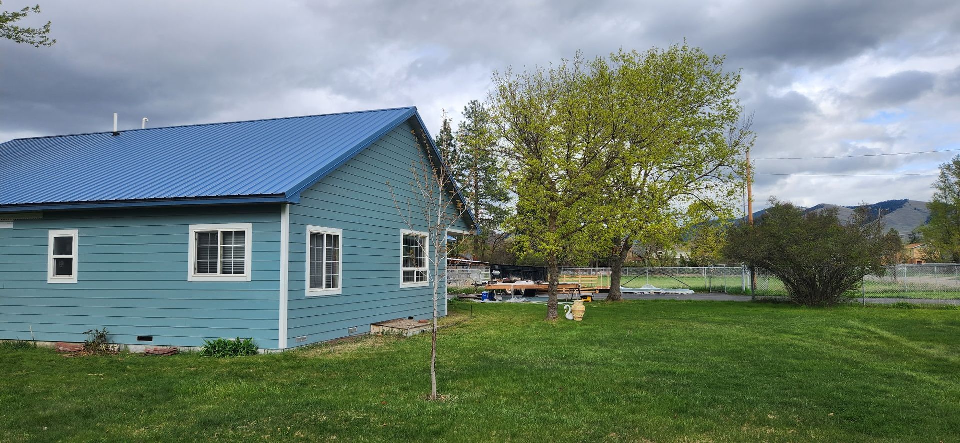 A blue house with a blue roof next to a tree, with green grass and an overcast sky.