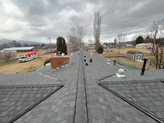 Rooftop with gray shingles, vents, and chimneys overlooking a rural landscape under cloudy skies