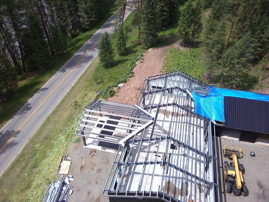 Construction site: metal framework of a building with a tarp-covered roof, next to a road and trees.