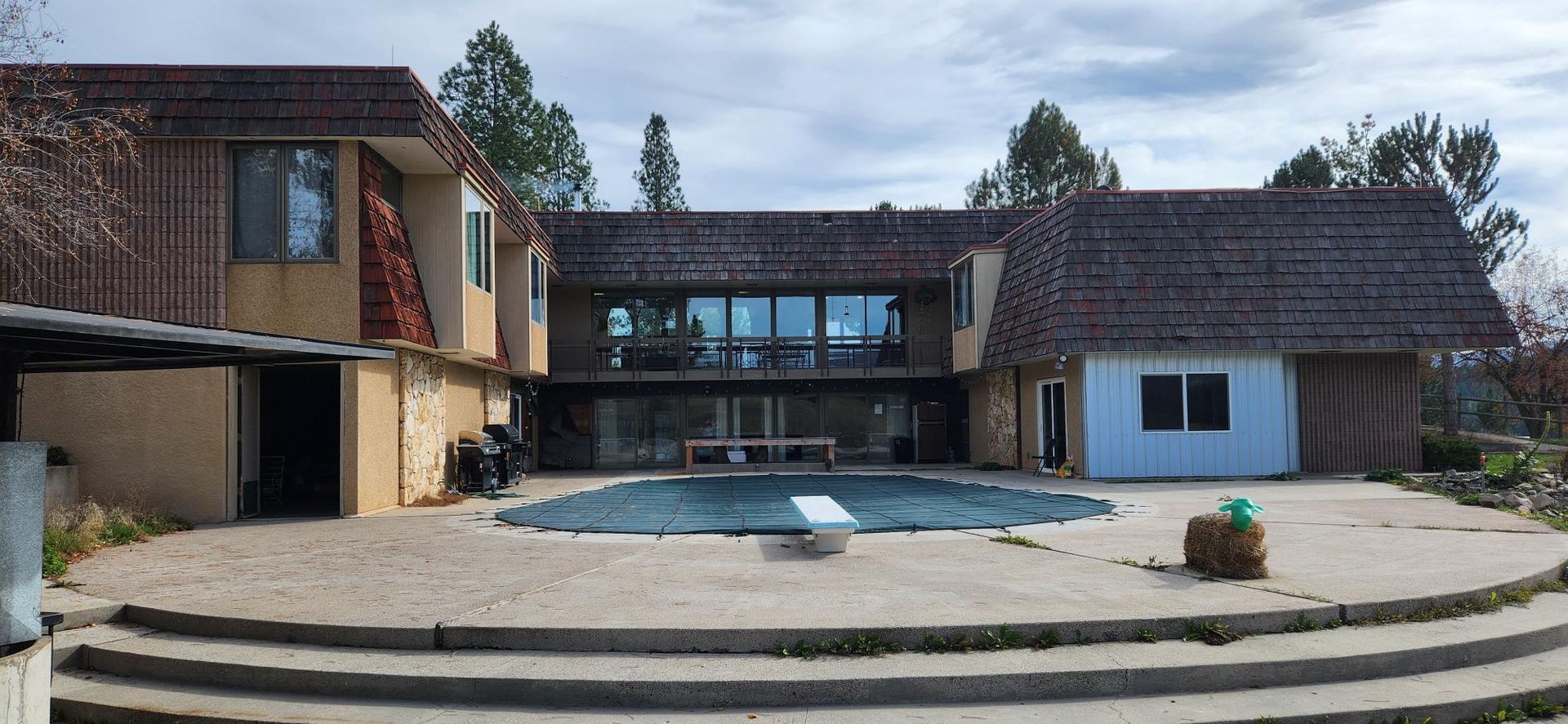 Backyard of a house with a pool, concrete patio, and trees under a cloudy sky.