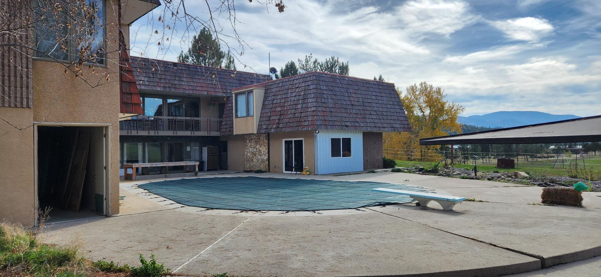 House with pool; cloudy sky. Stone-covered building, light blue painted section. Dive board, patio, and garage.