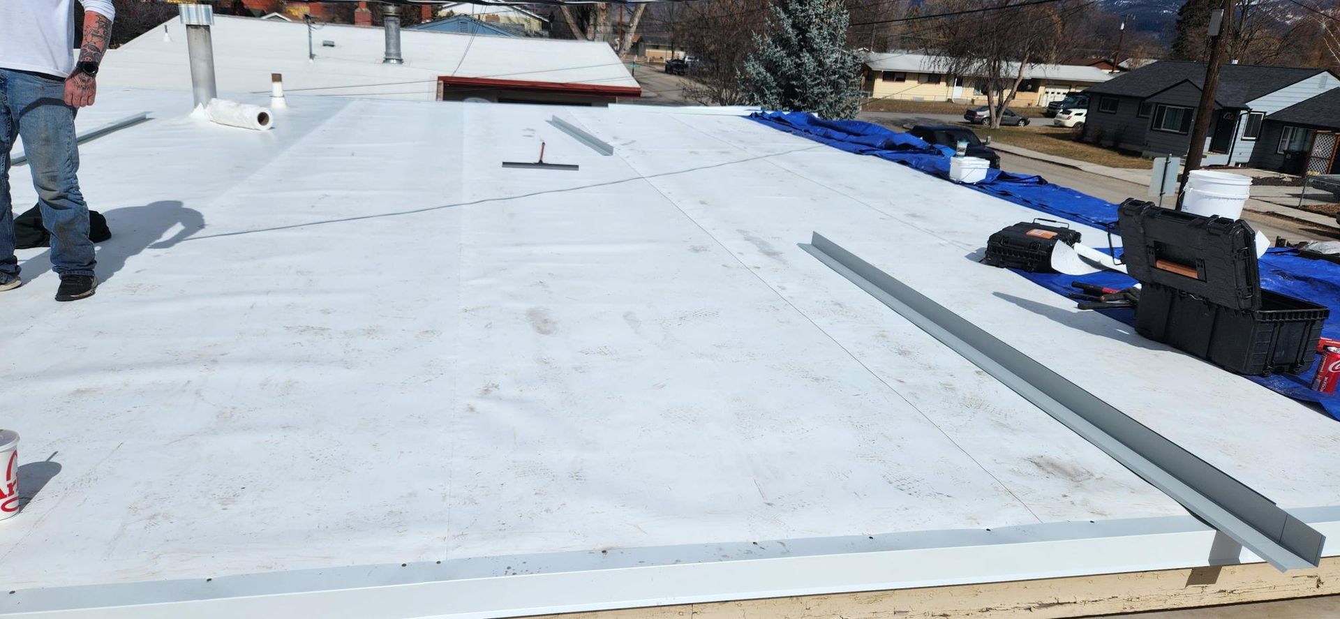 A person on a white roof with equipment. A metal piece is near the edge, blue tarps in the background.
