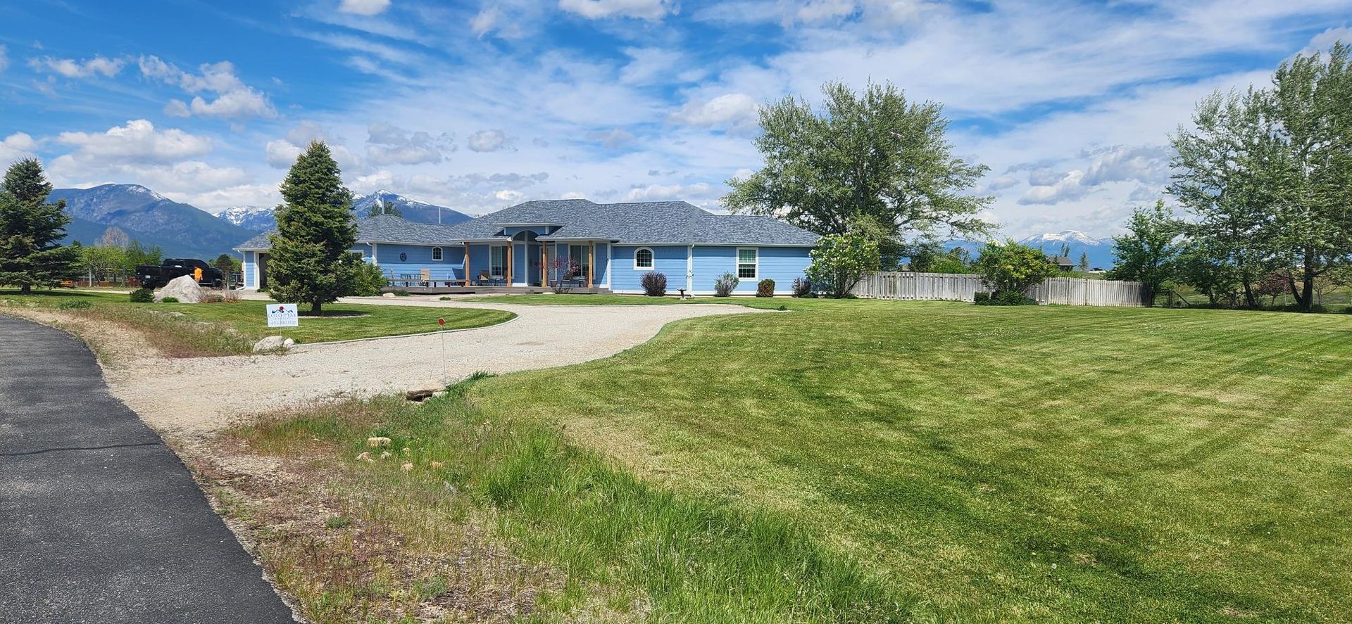 Blue house with gravel driveway and green lawn, mountains in the background, blue sky.