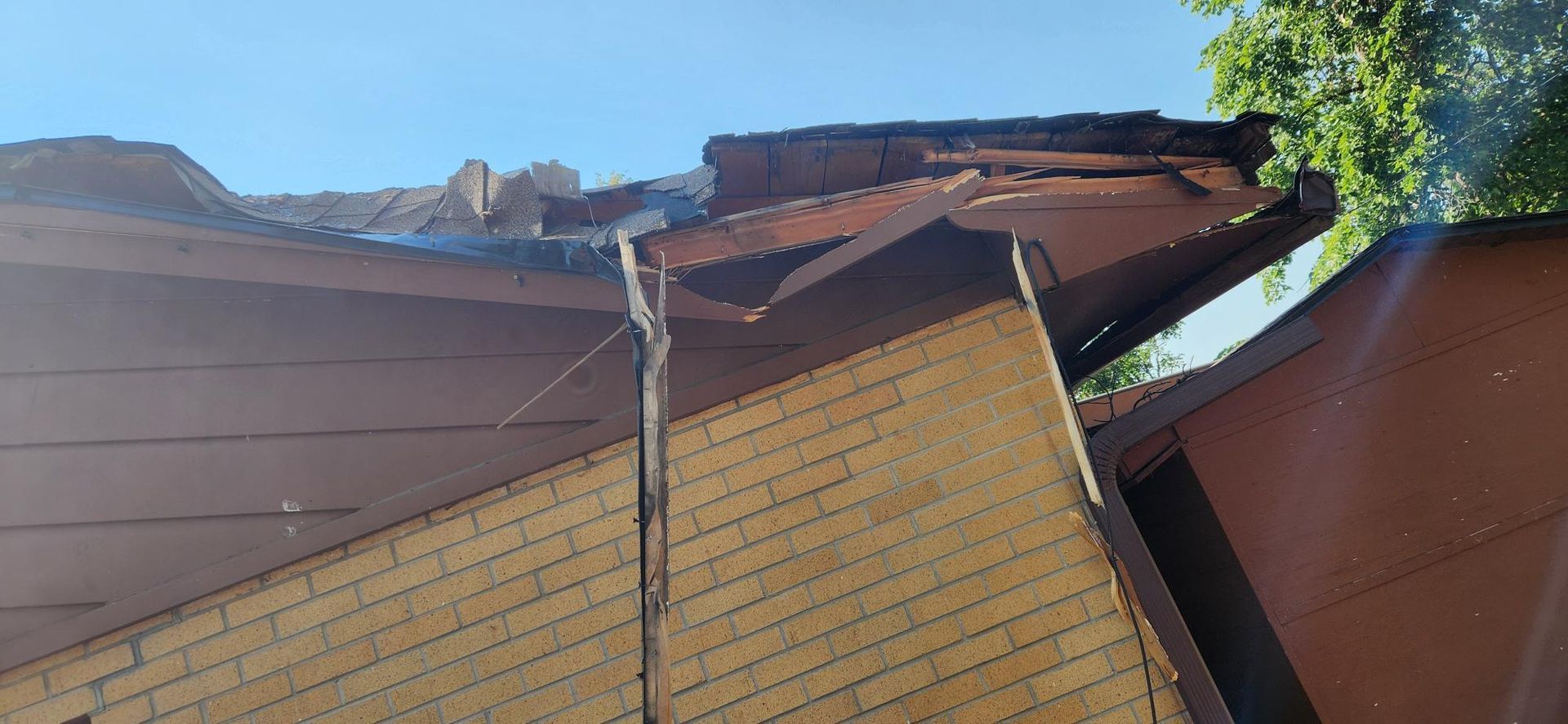 Damaged brown roof with missing shingles, angled against a blue sky, revealing roof structure.