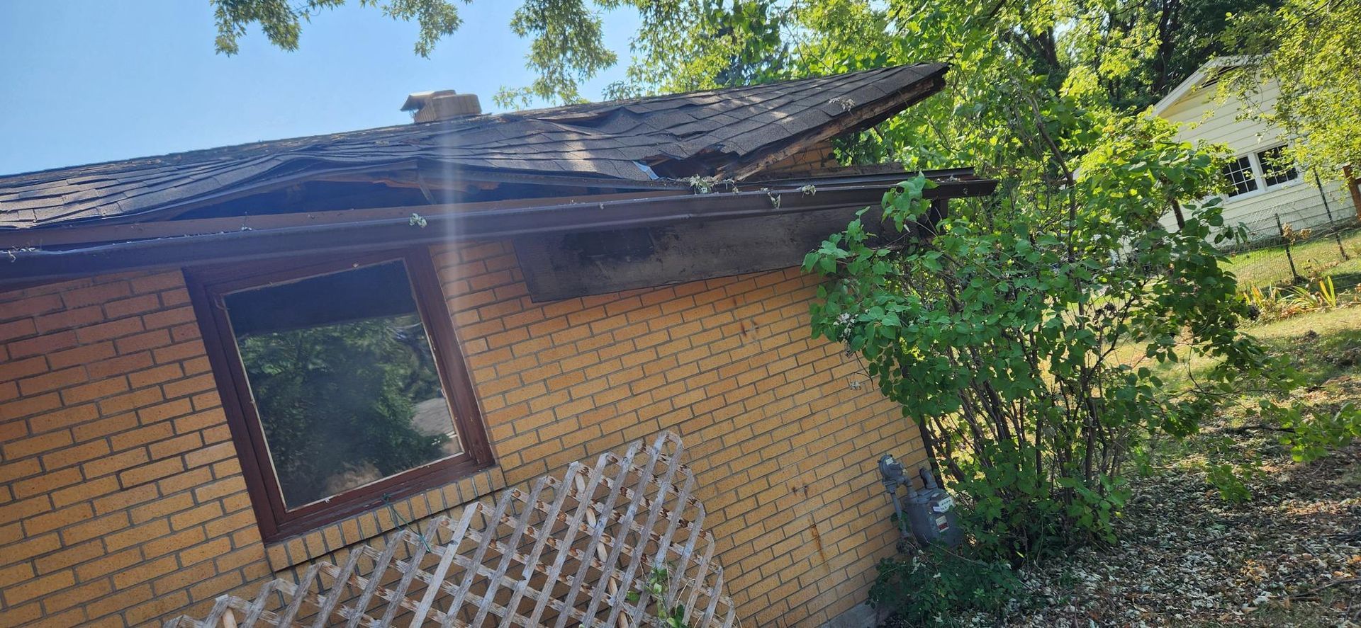 A brick building with a damaged roof, a window, and surrounding trees.