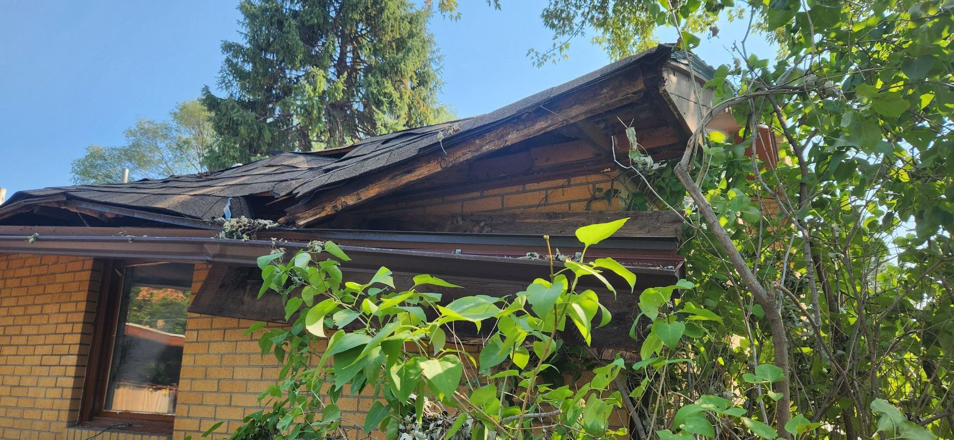 A damaged house roof with missing shingles. Brick exterior, green foliage, and a blue sky in the background.