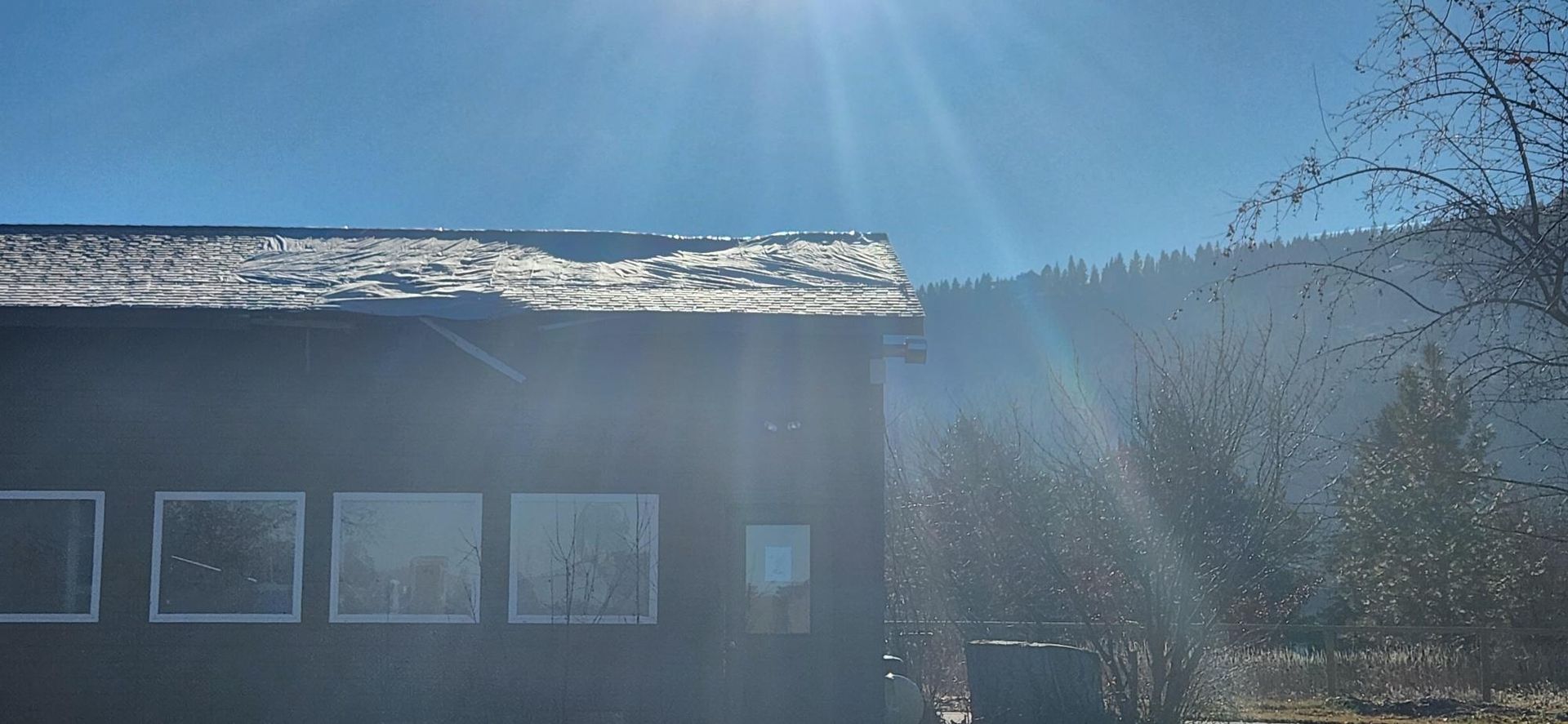 A sunlit wooden cabin with melting snow on the roof, trees in the background, blue sky.
