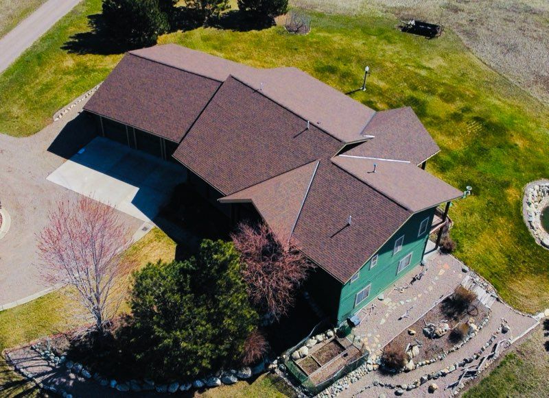 Aerial view of a brown-roofed green house with driveway, lawn, and trees on a sunny day