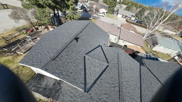 Aerial view of a gray shingled roof with vents and nearby trees and houses.