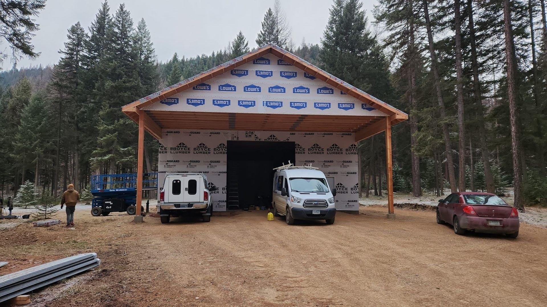 A partially-built wooden shed with vehicles parked in front, set in a wooded area.