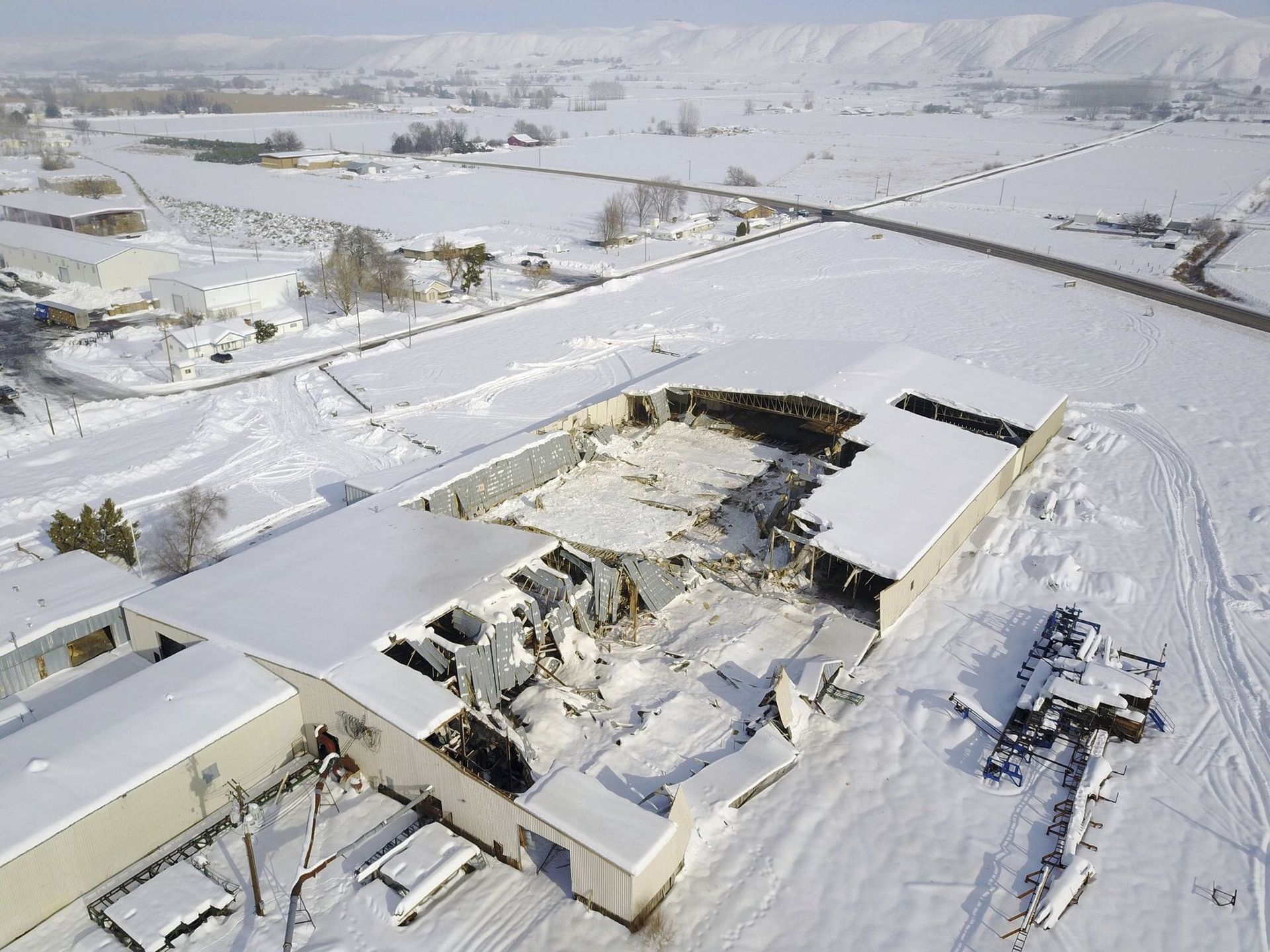 Snow-covered building with collapsed roof, aerial view. Snowy field and road in the background.