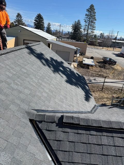 Roof shingles and a corner seam on a house, with a worker in orange visible at the edge and a neighborhood in back