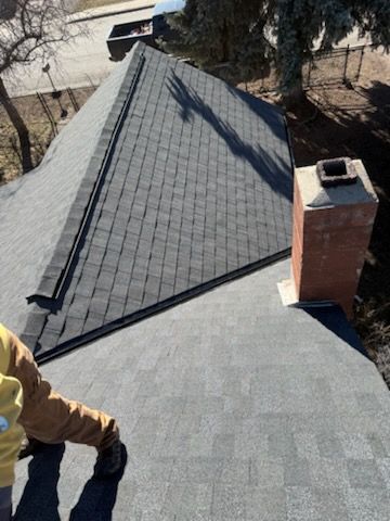 Roof under construction with a worker in a tan jacket, black shingles, and a brick chimney in view
