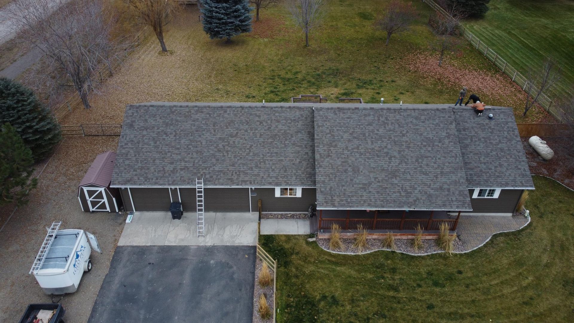 Aerial view of a brown house with a dark gray roof. A ladder leans against the side. A work truck is parked on the driveway.