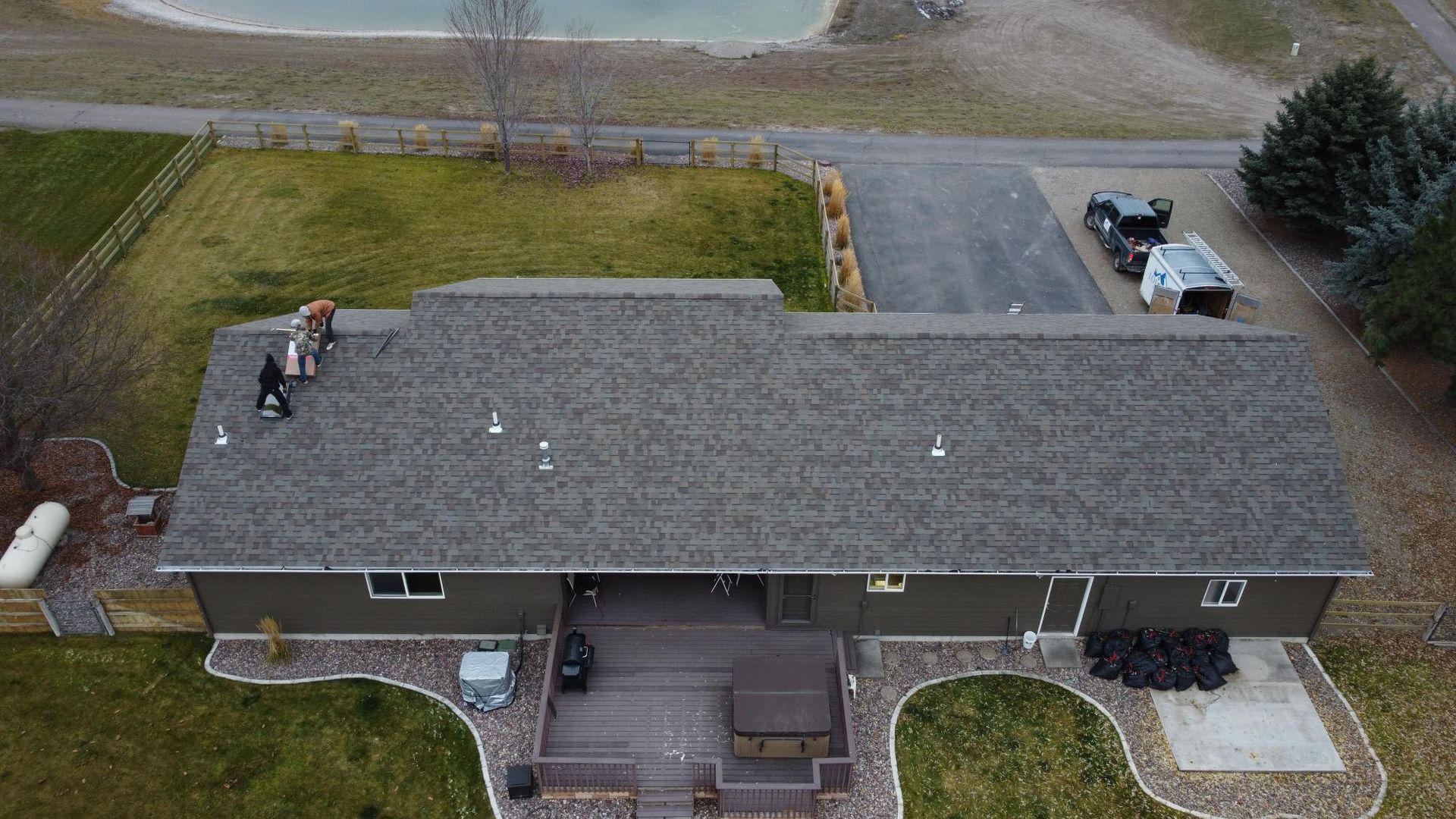 Overhead view of a house with a person on the roof. Dark gray roof and siding, brown deck, and grassy yard.