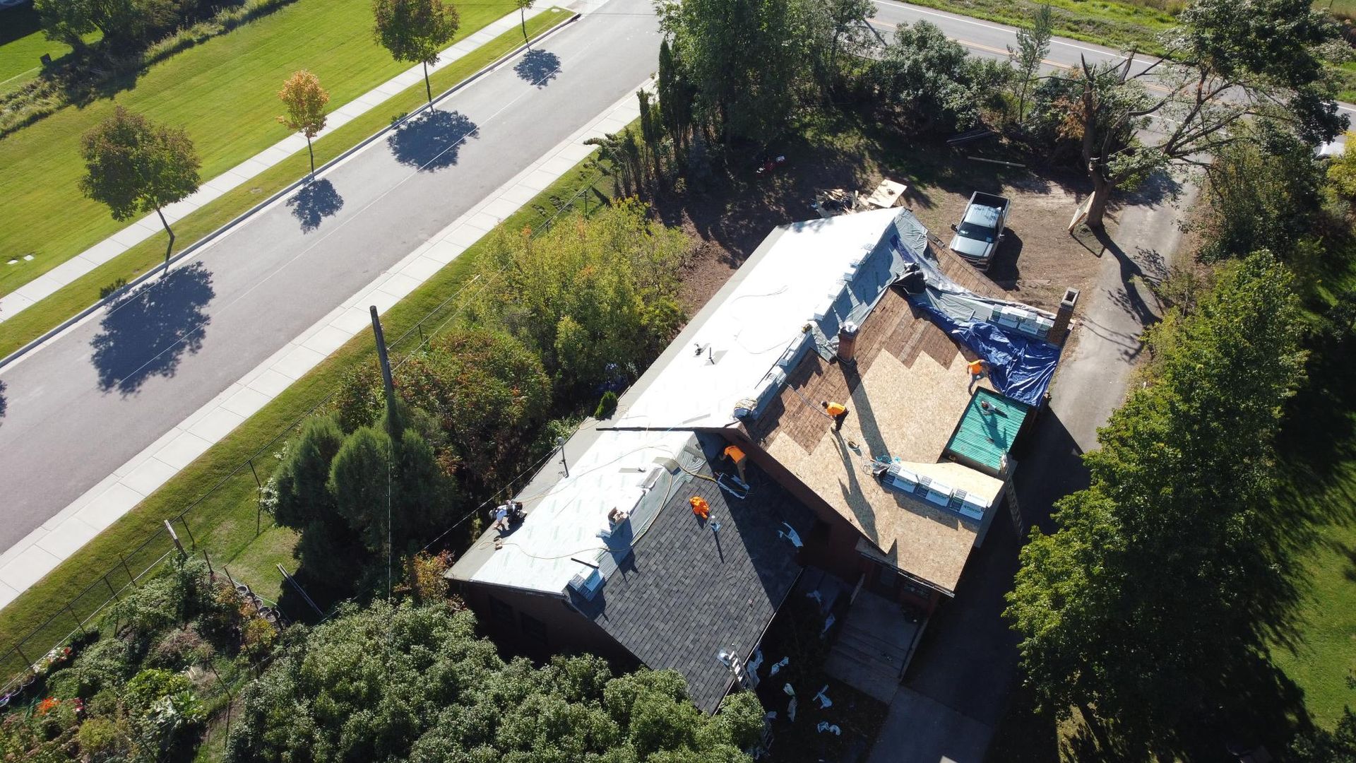 Overhead view of a house with roof replacement in progress. Workers on roof, blue tarp, asphalt shingles, and trees.