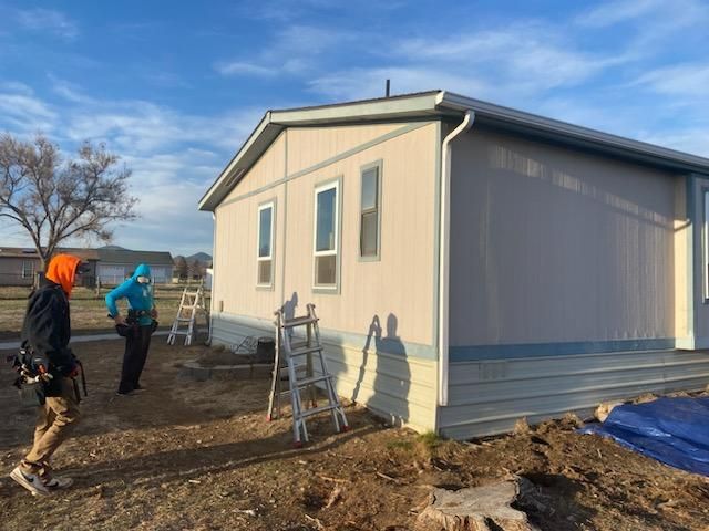 Three people near a light beige mobile home on a sunny day. Ladders are leaning against the side.