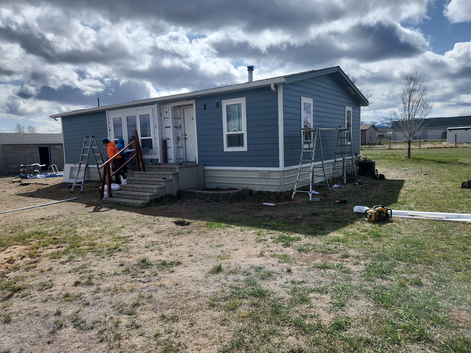 Blue siding home under construction; workers on porch, ladders visible; cloudy sky.