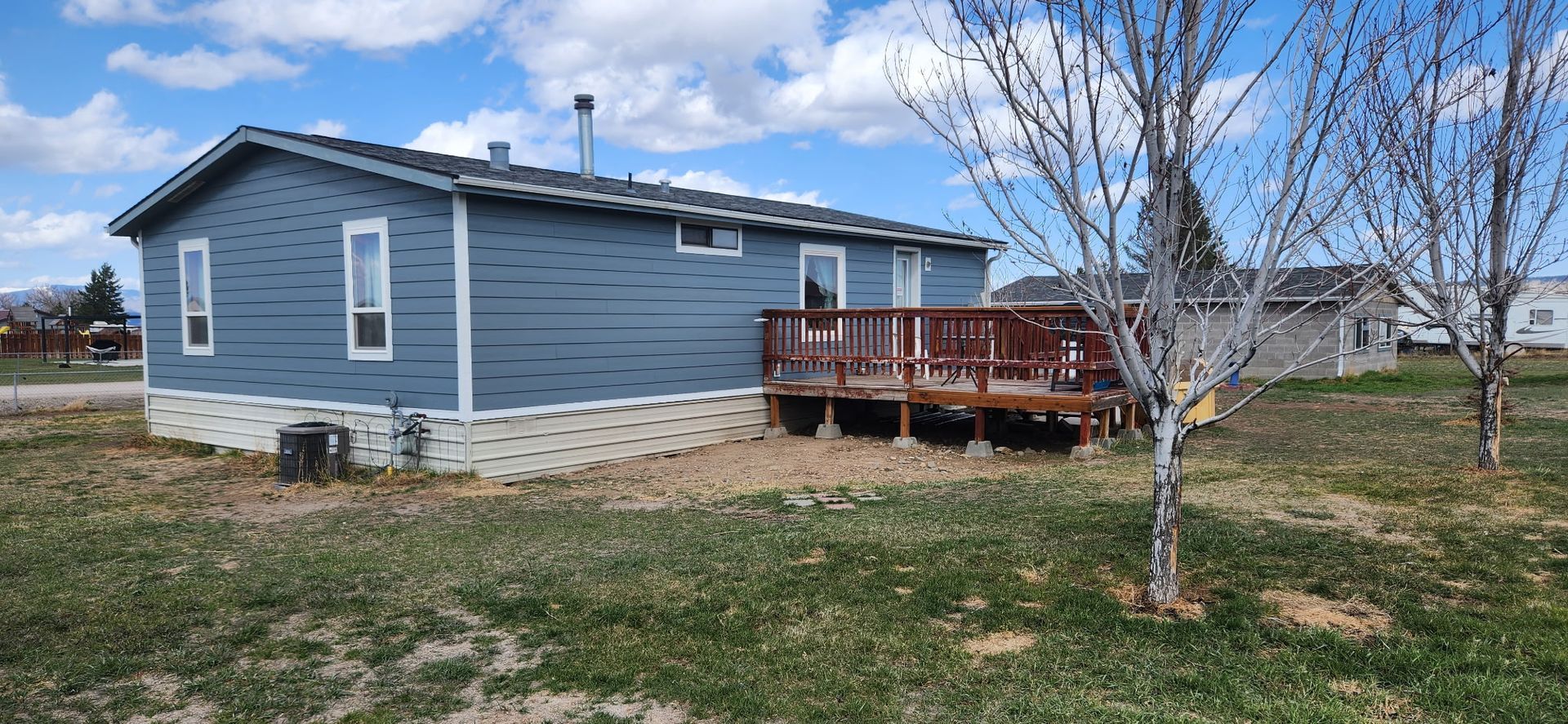 Blue-sided house with a wooden deck; a tree with no leaves in front of the house, grass and sky.
