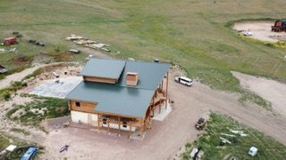 Aerial view of a small wooden house with a teal roof in a grassy rural area, with a parked vehicle nearby.