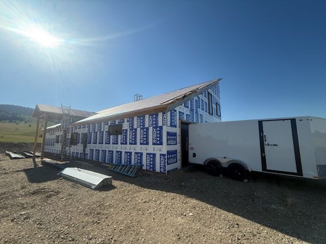 Partially built house wrapped in white-and-blue sheathing, with a trailer beside it under a bright blue sky