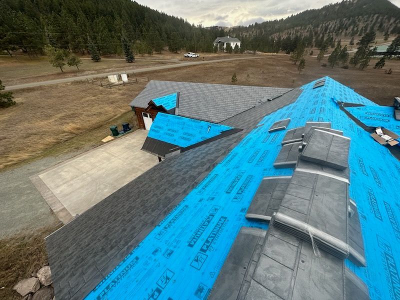 Roof under construction with blue underlayment and gray shingles on a house in a wooded mountain area