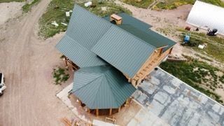 Aerial view of a green-roofed wooden building beside a paved area and dirt lot