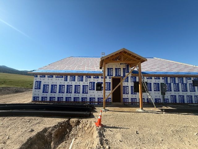 Small house under construction with blue-and-white wrapped walls and a wooden porch in a sunny rural area
